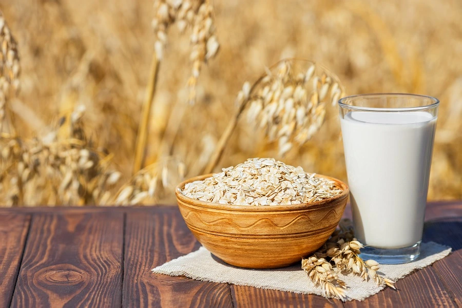 oat grain in wooden bowl next to glass of oat milk on brown table